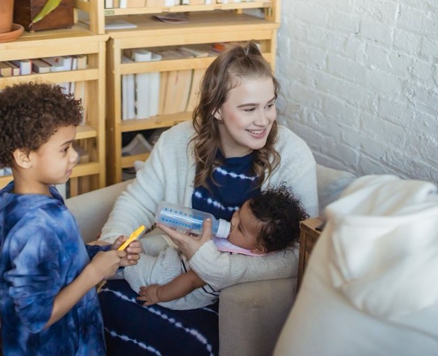 Au pair feeding a baby a bottle while a young boy sits beside them