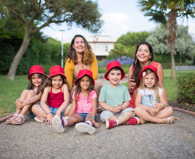 Two au pairs with a group of children in red caps in a park